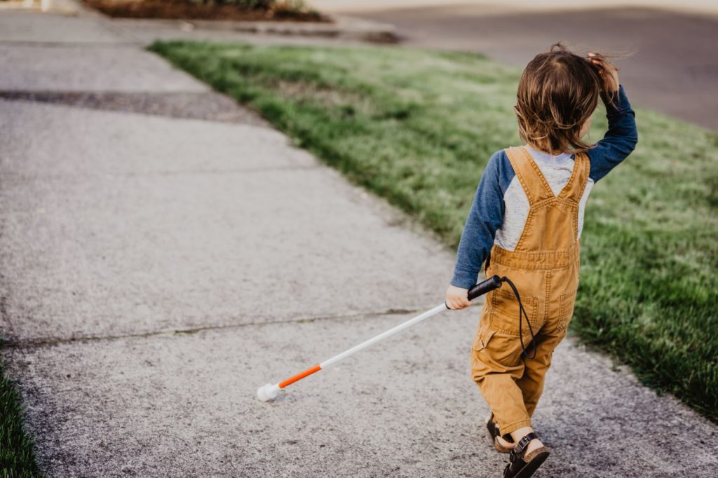 Vision impaired child in yellow dungarees walking along the pavement with a cane