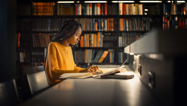 A picture of a woman in a yellow jumper writing notes on a laptop in a library, sitting in front of a bookshelf filled with books.