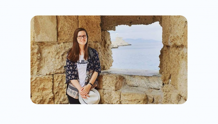 A picture of Fiona standing by a window of an ancient building, most probably a castle. She is looking at the camera and smiling, holding a hat in her hand.