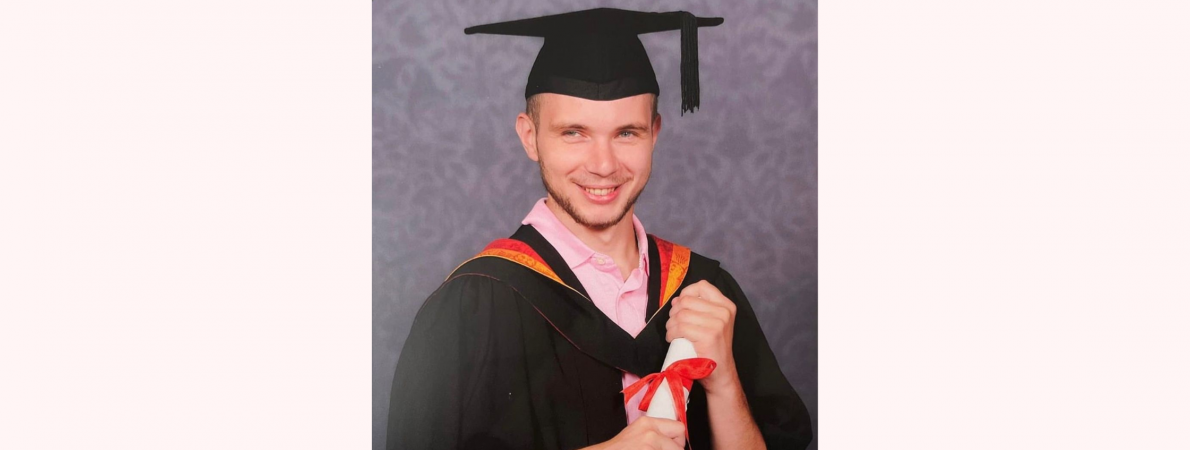 A headshot of Connah wearing his graduation hat and gown and holding his degree. He is smiling at the camera.