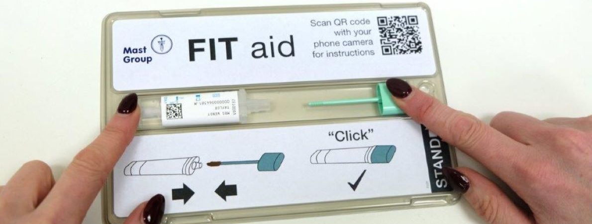 Image shows an open FIT aid test kit. The top right of the lid has a QR code, the top left depicts the logo of Mast. A woman's hands are sliding together the test tube, in her left hand, and the dip stick in her right hand, using the specially designed channel in the test box. The base of the test kit box depicts a graphic representation of sliding the two sections together.