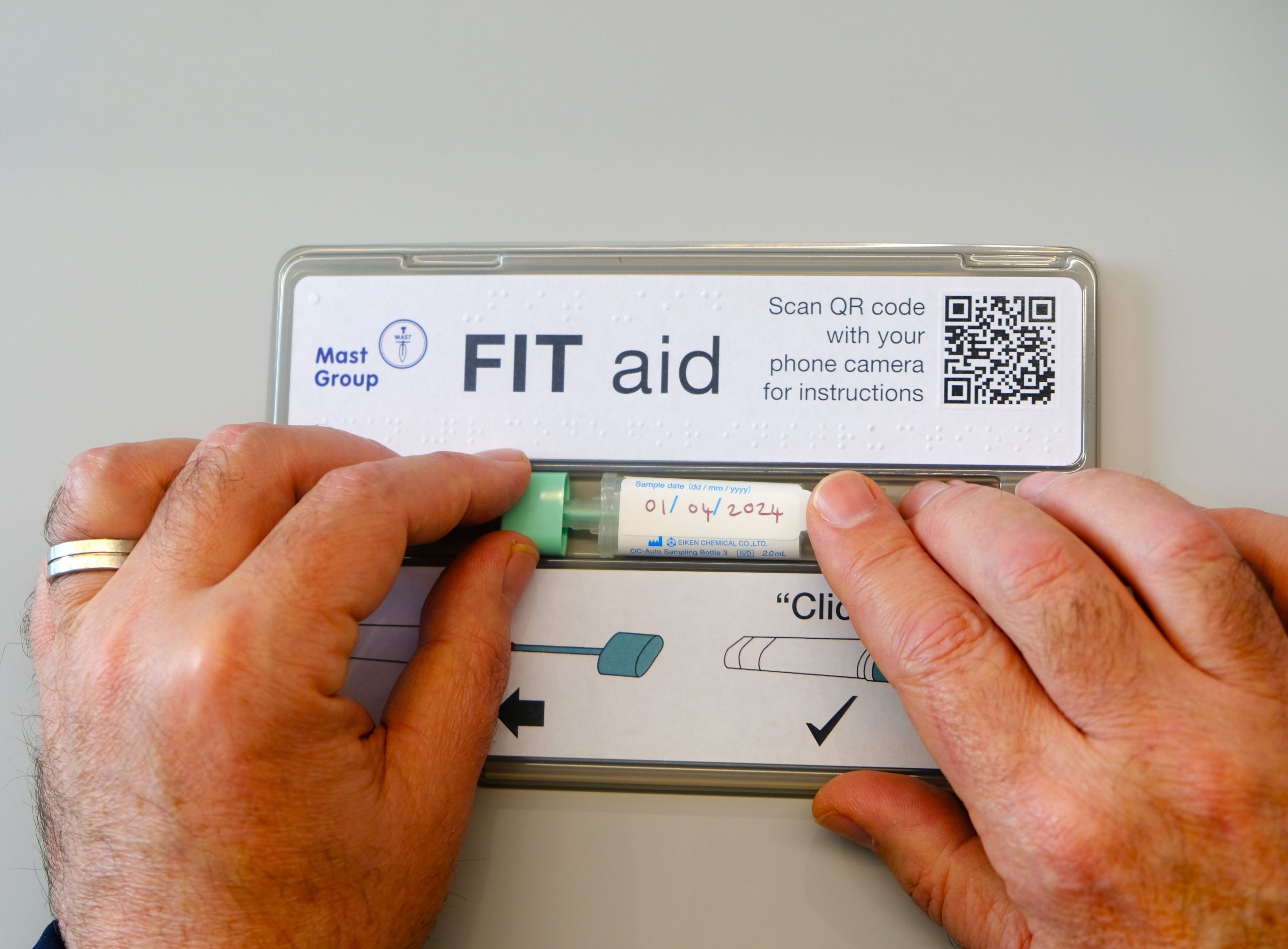 Image shows pair of hands using specially designed FIT aid tool. The open FIT aid box is placed on a surface. The pair of hands is sliding the dip stick and bottle towards one another, within the channel in the box, closing them together.