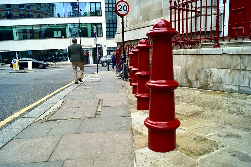 A London street seen from low level. There are red bollards along the right hand side of the pavement. In the distance a man walks close to the road next to a bicycle locked to railings.