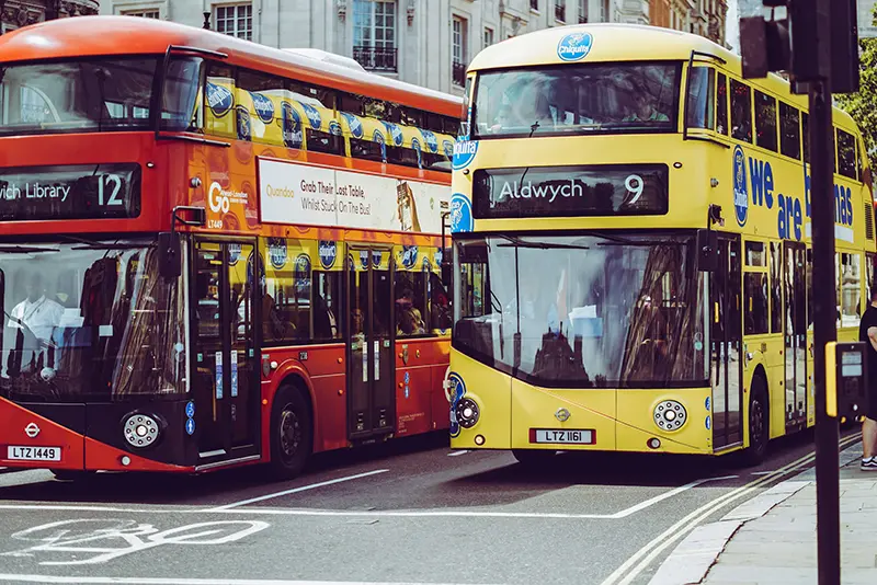 Two double-decker buses on a London street. The red bus is labeled number 12, and the yellow bus, advertising Chiquita Bananas, is labeled number 9 to Aldwych