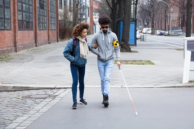 The image shows two individuals walking on a sidewalk. The person on the right is using a white cane, which suggests they may have a visual impairment. The person on the left is carrying a backpack and appears to be accompanying the individual with the white cane