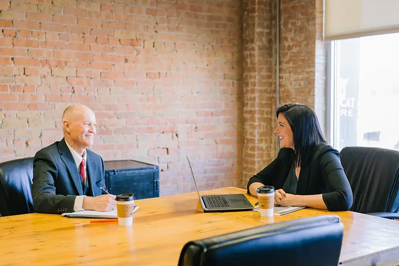 A smartly dressed lady sits at a meeting room table in front of a laptop, she is smiling across the table at man dressed in a suit. The man is making notes in a notebook.