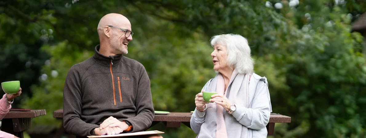 Two individuals chat on a park bench. One man is wearing shorts and an outdoors style fleece. He smiles as a lady with grey hair chats to him. She is holding a green cup of coffee.