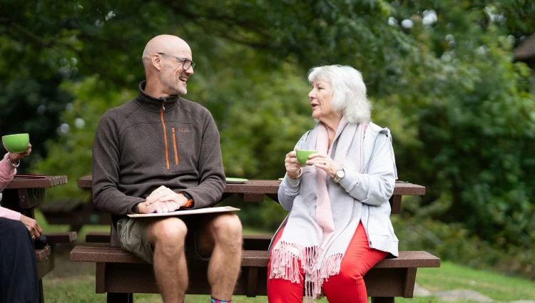 Two individuals chat on a park bench. One man is wearing shorts and an outdoors style fleece. He smiles as a lady with grey hair chats to him. She is holding a green cup of coffee.