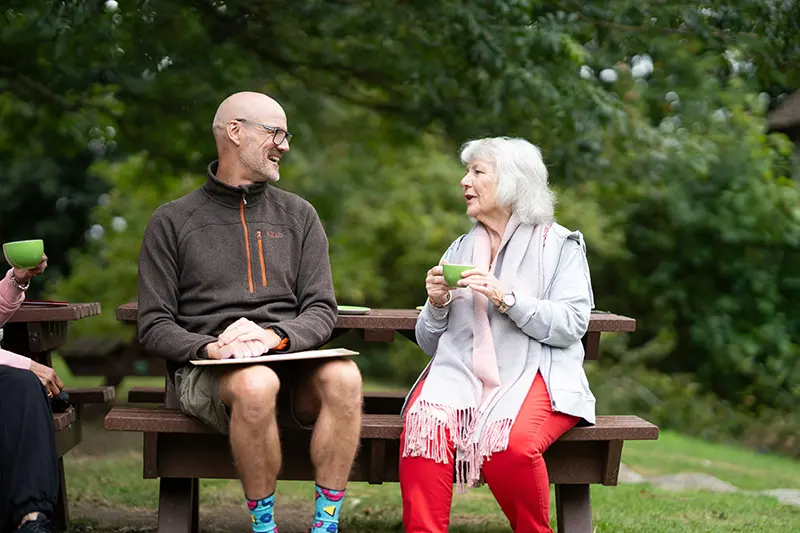 Two individuals chat on a park bench. One man is wearing shorts and an outdoors style fleece. He smiles as a lady with grey hair chats to him. She is holding a green cup of coffee.