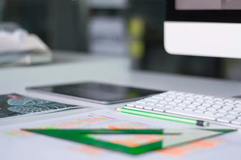 Image of a desktop. On the desk is a iMac computer with a white keyboard, a green pencil and some notes highlighted in orange.