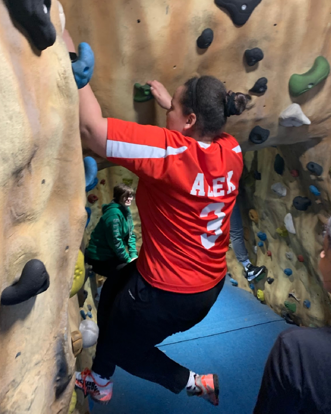 A picture of Alex climbing an indoor rock wall in a red shirt with her name on the back, and black trousers, having a great time.