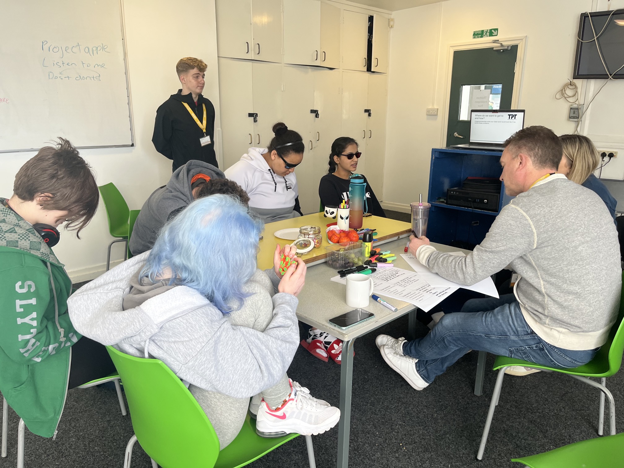 5 students sit around a table with two adults. One adult is stood in the background. A laptop is placed on some shelves. On the table is a range of paper, highlighters, water bottles and snacks. There is a whiteboard in the background. One adult is writing notes on paper.