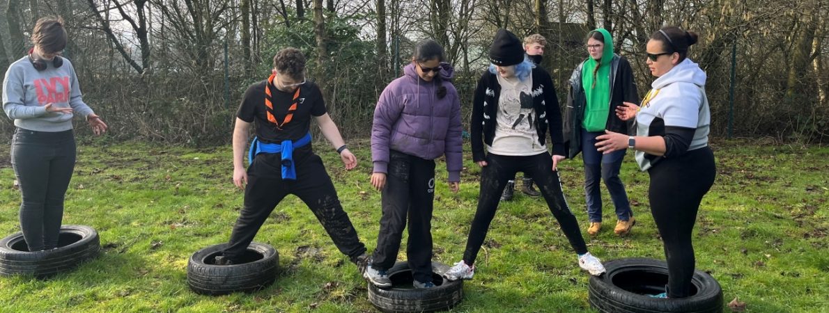 five students are spread across four tyres, balancing between them. There are two adults in the background. The students are communicating with each other and appear to be trying to solve a problem. They are on grass with trees in the background.