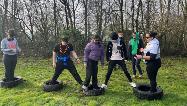 five students are spread across four tyres, balancing between them. There are two adults in the background. The students are communicating with each other and appear to be trying to solve a problem. They are on grass with trees in the background.