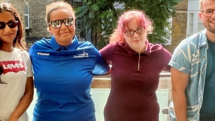 A picture of the Young Voices Sports team smiling at the camera, standing side by side in front of a glass railing wit a building and trees in the background.