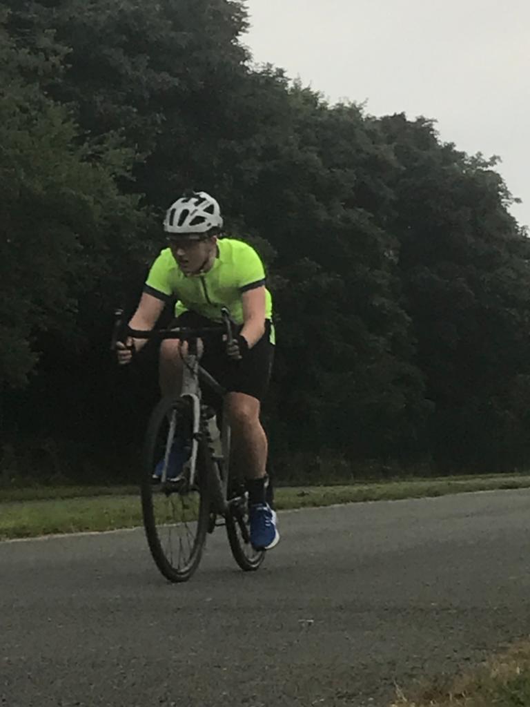 A picture of Tom riding his bike outdoors on a paved path, wearing a yellow shirt and black shorts, looking super concentrated as he pedals by some trees.