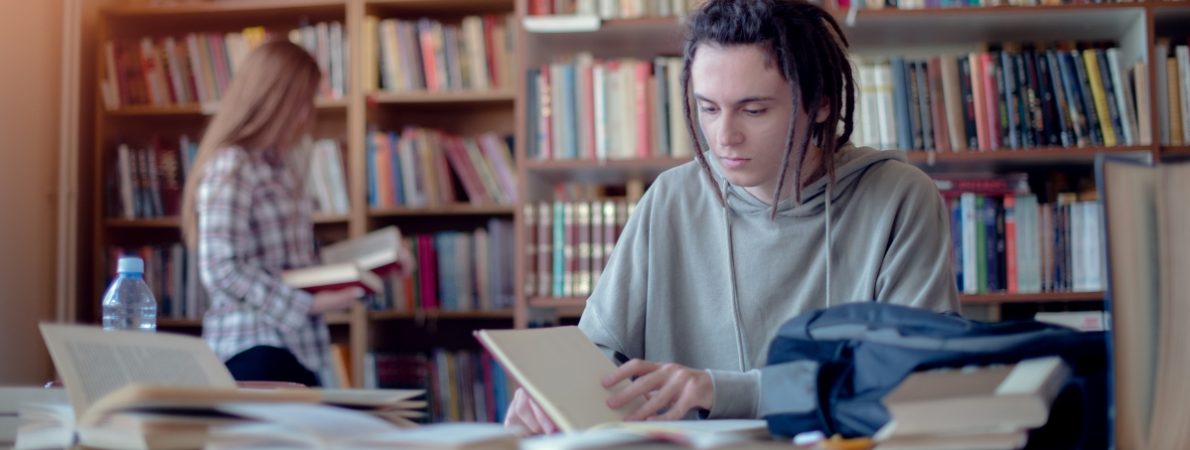 Young student learning in library, female student choosing books from bookshelf in background.