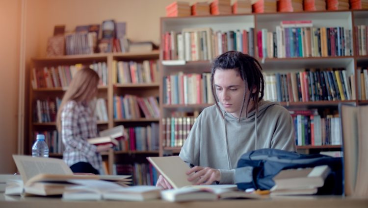 Young student learning in library, female student choosing books from bookshelf in background.