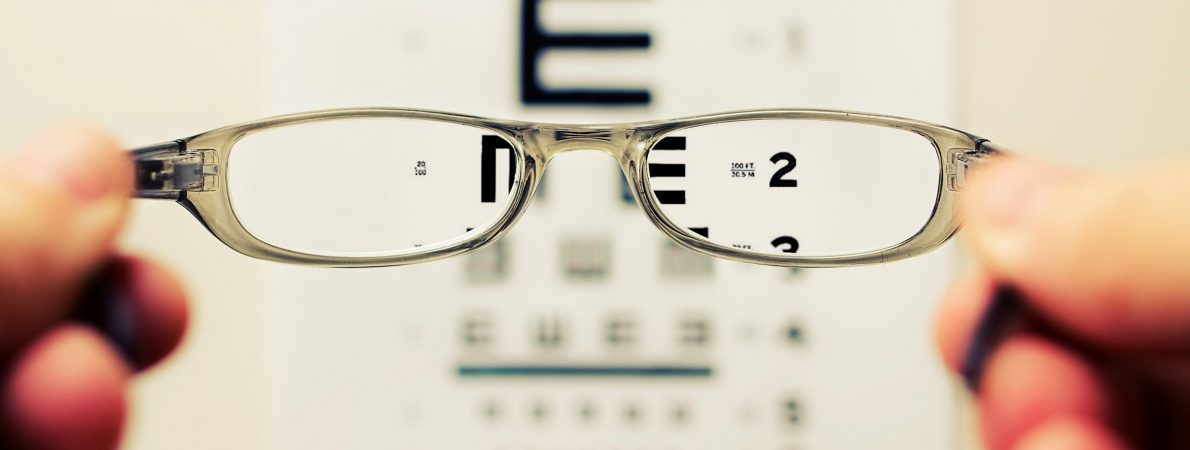 Photograph of an eye test chart being viewed through the aspect of a pair of sliver, metal rimmed glasses. The image through the lenses is clear, but otherwise blurred.