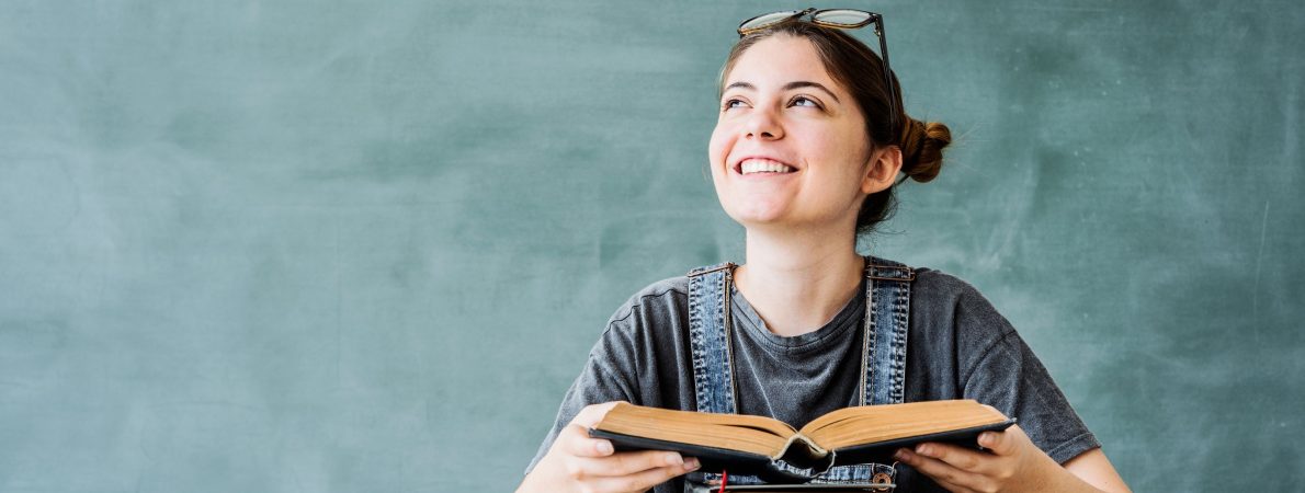 young student with a pile of books, holding a book, smiling and looking up