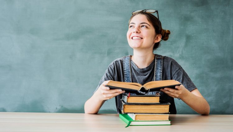 young student with a pile of books, holding a book, smiling and looking up