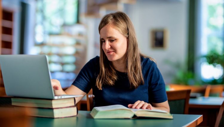 A visually impaired woman sitting and studying in the university library using a laptop and books.