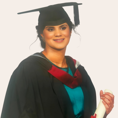 A graduation photo of Nayab, a South Asian woman, with an updo hairstyle and subtle, but beautiful natural make up. Nayab is wearing a black mortarboard hat with the tassel at the front, a black graduation gown with a red satin ribbon. The gown is draped over a teal coloured dress. Nayab holds a rolled up, cream diploma tied with a red ribbon.