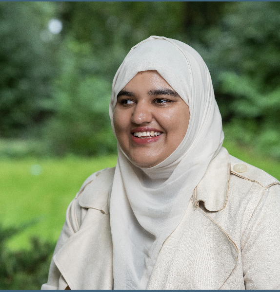 A picture of Khansa smiling at the camera, wearing a light coloured hijab and matching coat, standing in a green outdoor space.