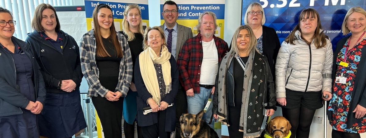 Picture shows a group of SLC volunteers standing alongside two Birmingham Community Healthcare workers. Two support dogs sit set at the front, held on leads by a partially sighted male and female. Two NHS roll up banners are displayed behind the group.