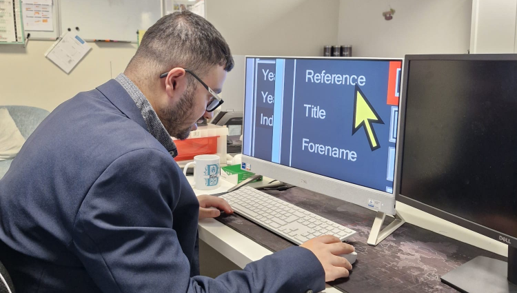 Bilaal, a young man with dark hair and sporting a beard, is smartly dressed in a suit and sat at his workstation in front of a computer