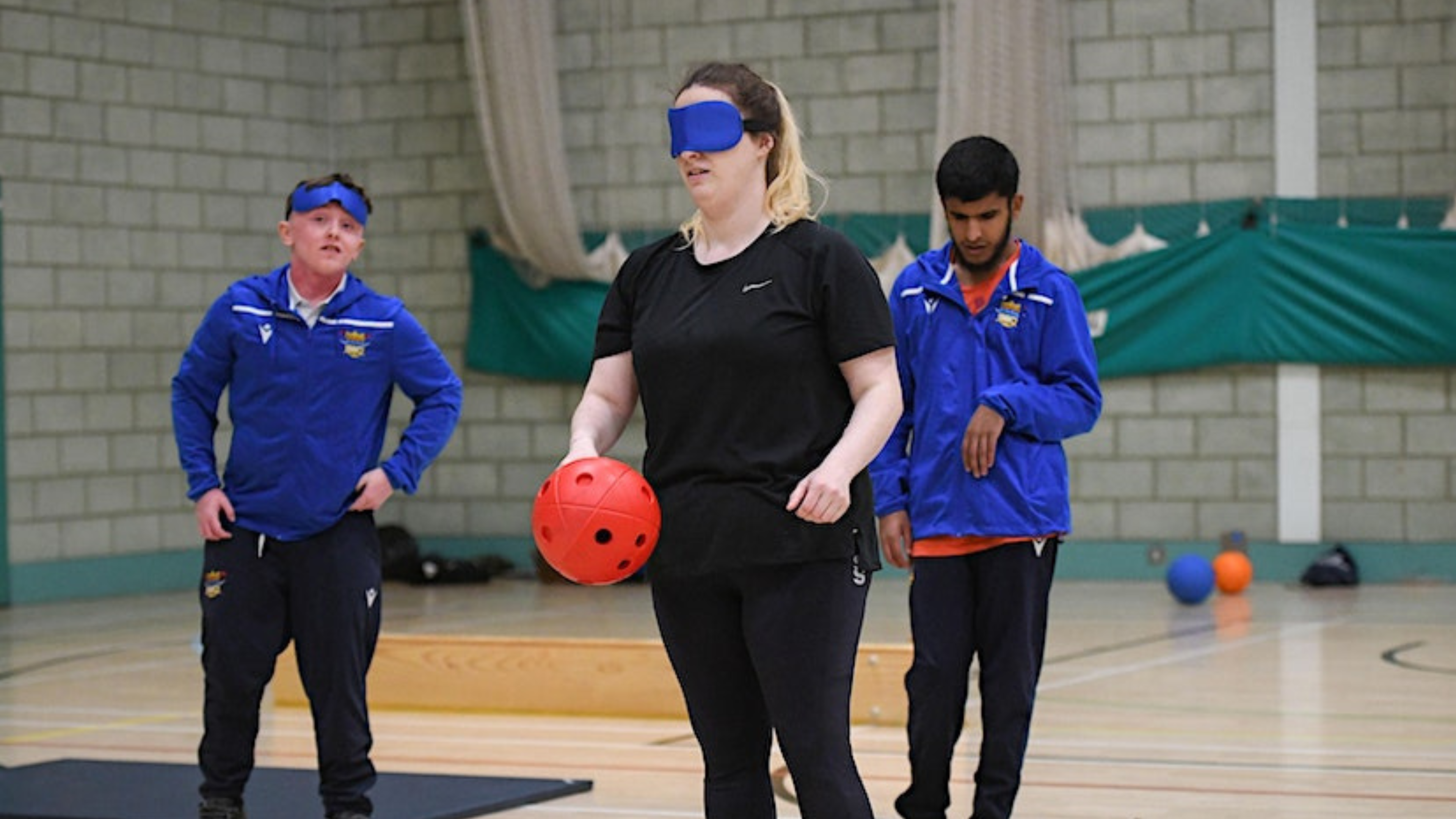 in a sports hall on a marked court, a woman holds a red adapted sports ball. The woman is wearing a blindfold. Behind the woman are two sports professionals looking towards the goal the woman is aiming for of camera. 