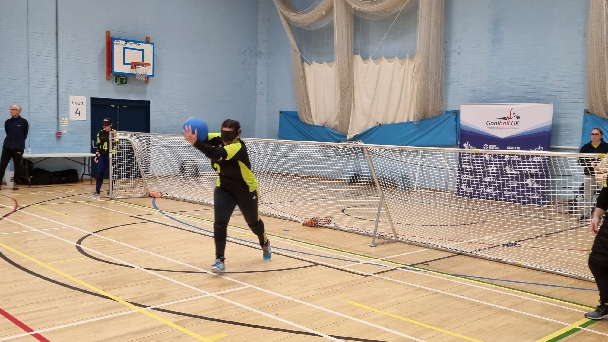 Young Voices Volunteer Leader Alex holds a goalball at head height in a position suggesting she is about to bowl the ball at speed towards the opposing team. Alex is wearing a blindfold and wears team colours of black and tennis ball yellow. Alex is stood in front of her team’s goal and is in an indoor sports hall with lines marking the court on the ground. Another member of her team can be seen to the side of the goal, and there is a sighted professional off-court.