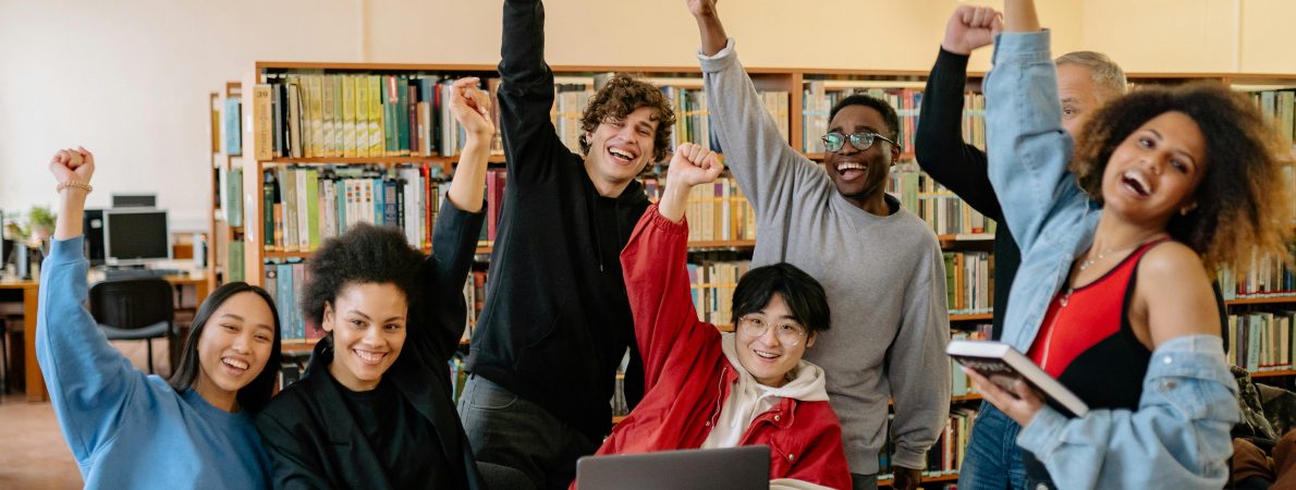 A diverse group of young people smiling with hands punching the air, seated and standing in a university library with books and laptops open on the table in front of them.