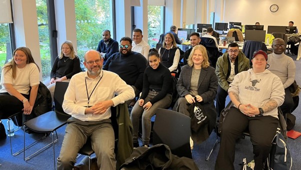 Photo of latest of interns 2024 to 2025 cohort: A group of interns of mixed genders, age, and ethnicities sit in an open office environment with large windows that allow plenty of daylight.  