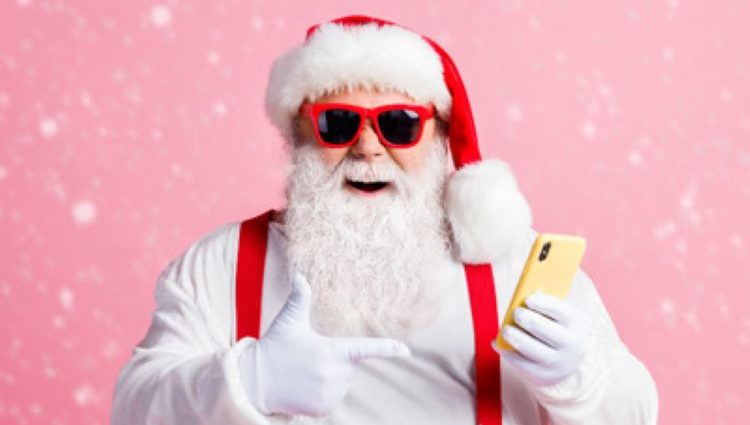 Santa points to his mobile phone in his left hand. He's forgotten his cloak so we're exposed to his red braces holding up his red trousers over a white shirt. He's wearing red rimmed sunglasses, his trademark hat and is clearly enjoying being photographed on a sparkly pink background.