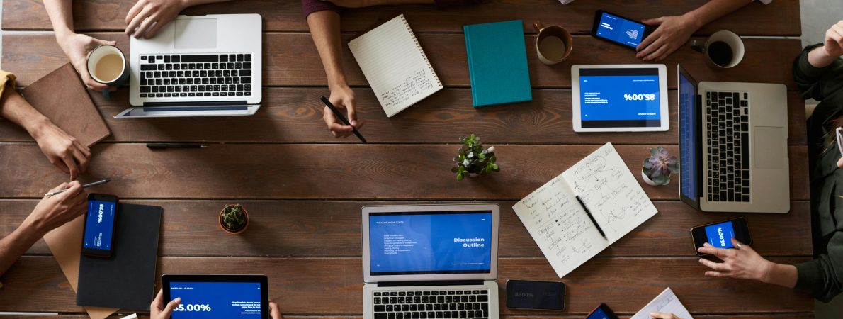 A picture of a collaborative work space with several people seated around a wooden table, each with laptops, tablets or notebooks in front of them, with coffee cups at the ready to take action.