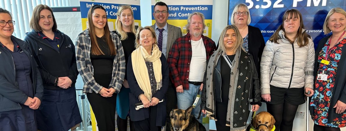 Picture shows a group of SLC volunteers standing alongside two Birmingham Community Healthcare workers. Two support dogs sit set at the front, held on leads by a partially sighted male and female. Two NHS roll up banners are displayed behind the group.