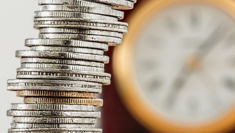 The picture shows a stack of various coins with a clock on the wall in the background