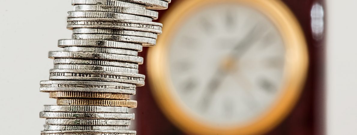 The picture shows a stack of various coins with a clock on the wall in the background