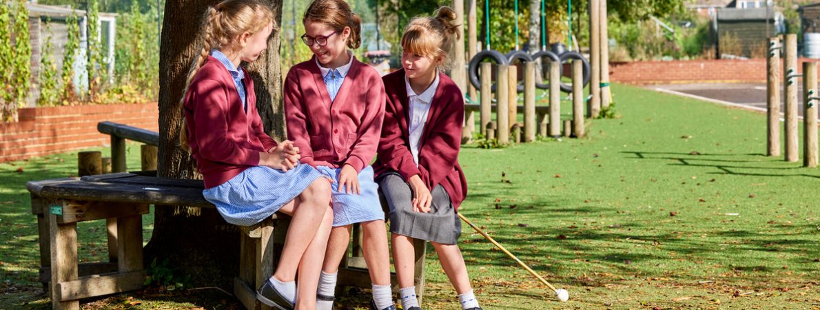 Three primary school aged children sit by a tree in their school uniform. Two are wearing a blue dress with a maroon cardigan over the top whilst the third is wearing a white blouse, grey skirt, the same maroon cardigan and is also holding a white cane. A playground is seen in the background.