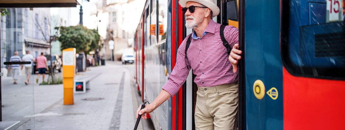 A blind or partially sighted older man uses his cane to step off a red bus