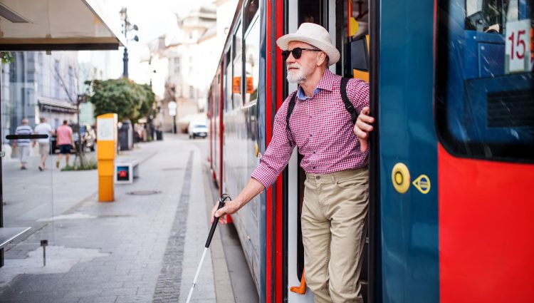 A blind or partially sighted older man uses his cane to step off a red bus