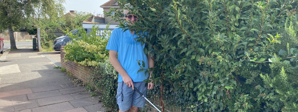 A man holding a white cane is impeded by an overgrown bush protruding from a front garden onto the pavement. He is wearing a blue polo, grey shorts and dark glasses.