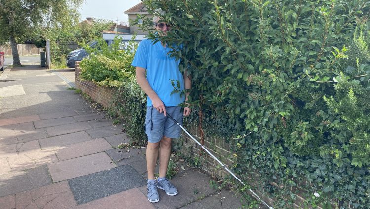 A man holding a white cane is impeded by an overgrown bush protruding from a front garden onto the pavement. He is wearing a blue polo, grey shorts and dark glasses.